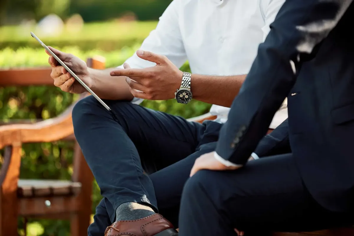 Zwei Personen sitzen draußen und besprechen etwas, eine hält ein Tablet und gestikuliert.