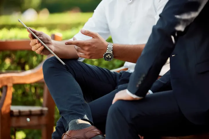 Zwei Personen sitzen draußen und besprechen etwas, eine hält ein Tablet und gestikuliert.